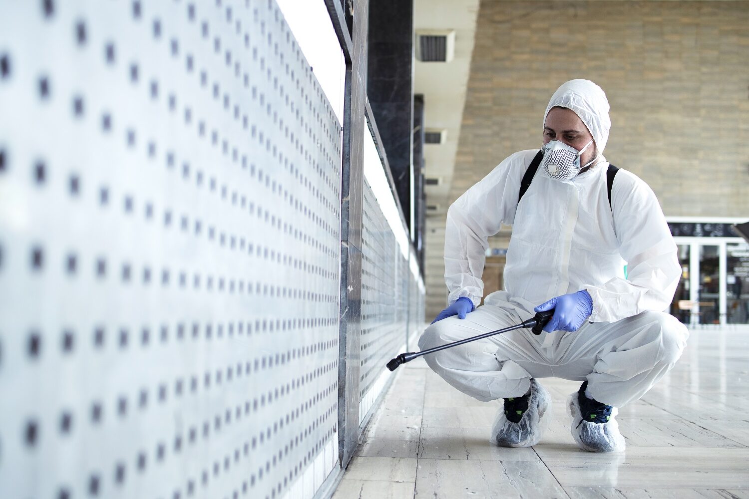 Unrecognizable worker in chemical protection suit spraying disinfectant on public surfaces.